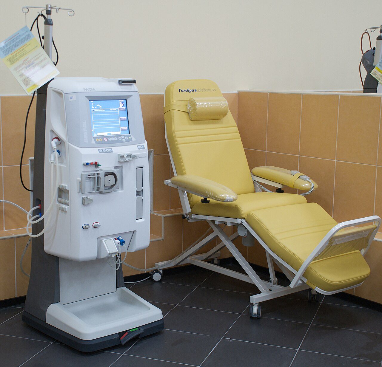 A white dialysis machine with a screen and tubing stands beside a yellow reclining treatment chair in a tiled clinic room