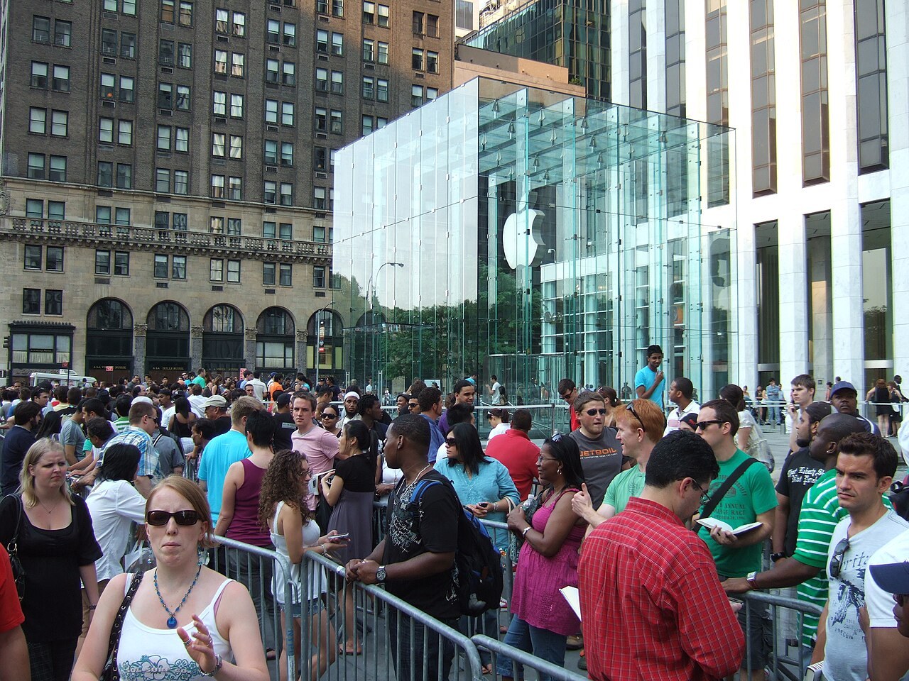 A crowd of people standing behind metal barriers in a long line outside a glass cube-shaped Apple Store entrance on a city plaza