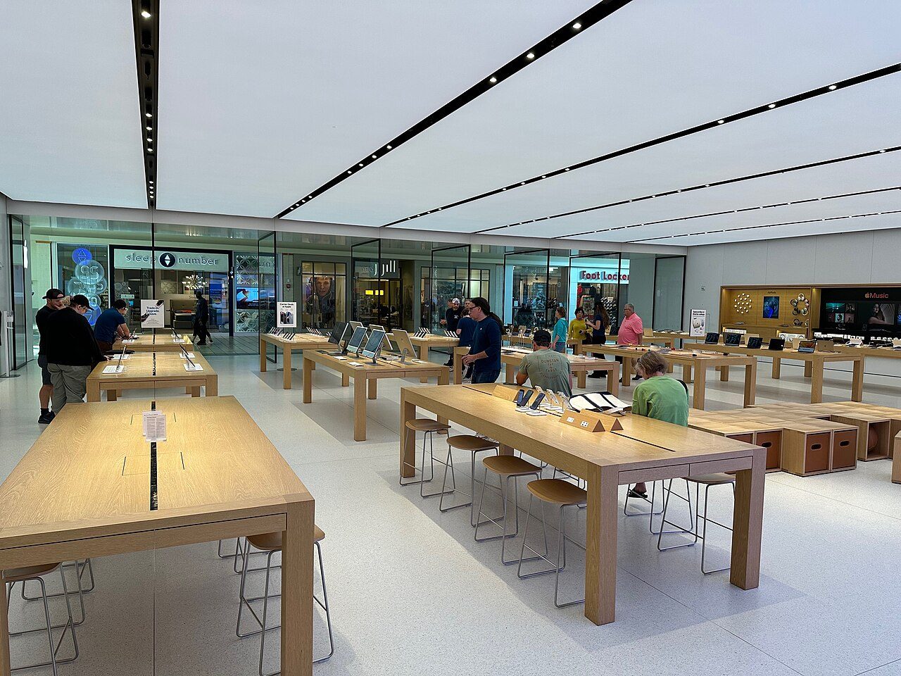 The bright interior of an Apple Store with long light wood tables of devices, white walls and shoppers browsing