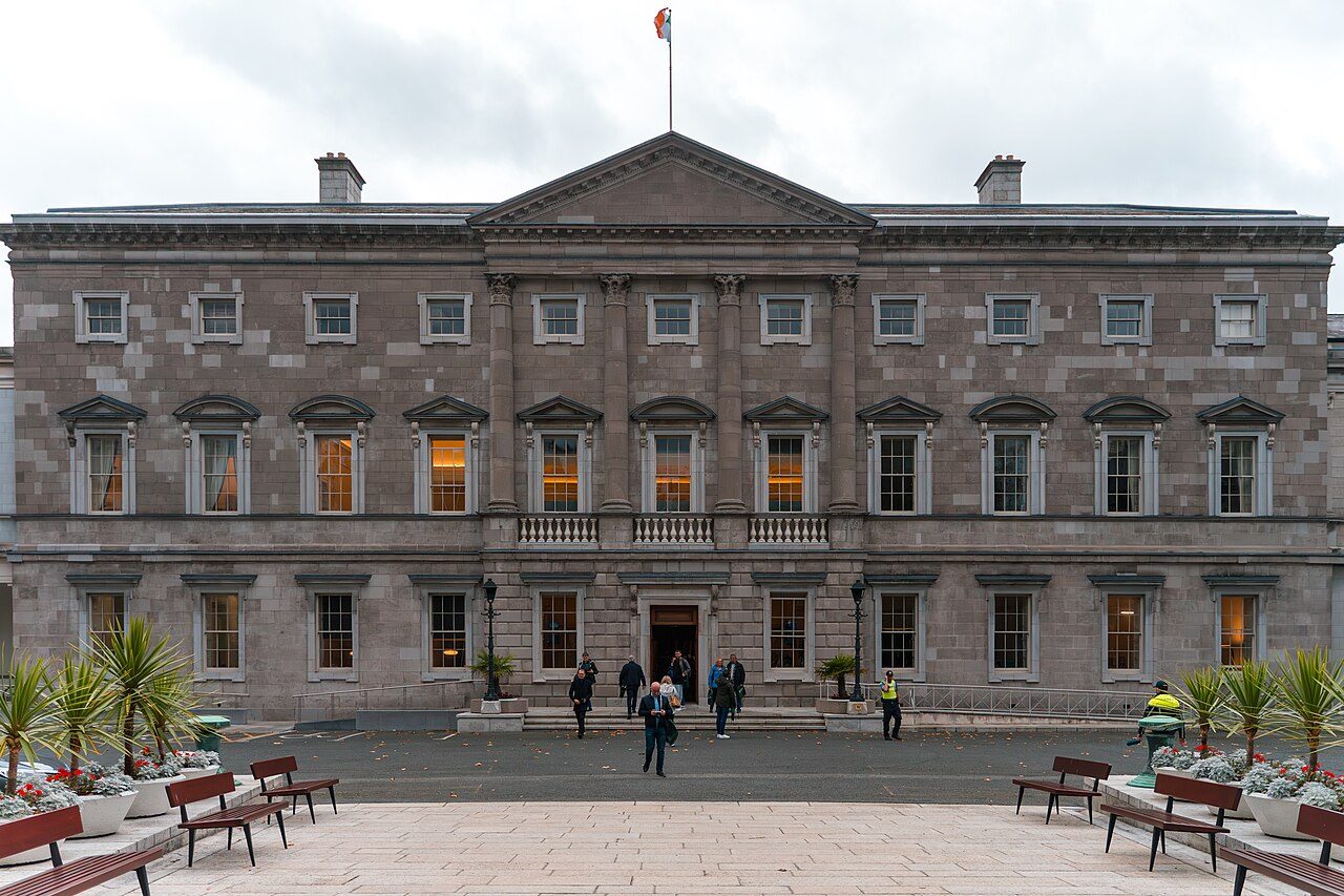 The gray stone front of Leinster House with a flag on the roof and benches in the courtyard