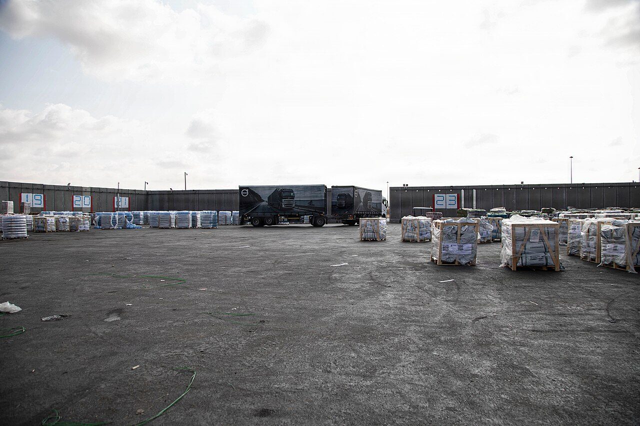 Pallets of wrapped boxes sit in rows on a wide paved lot inside a walled crossing area, with a truck parked near the far wall under a gray sky