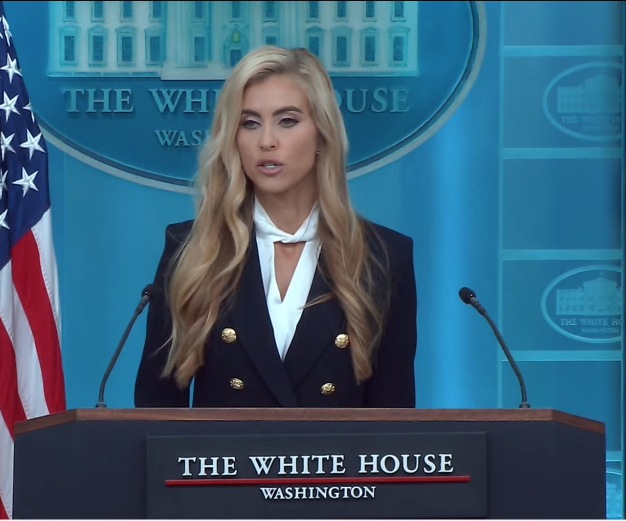 Katie Zacharia, with long blond hair, a dark double-breasted jacket with gold buttons and a white blouse, speaks at the White House lectern in front of a U.S. flag and the White House seal