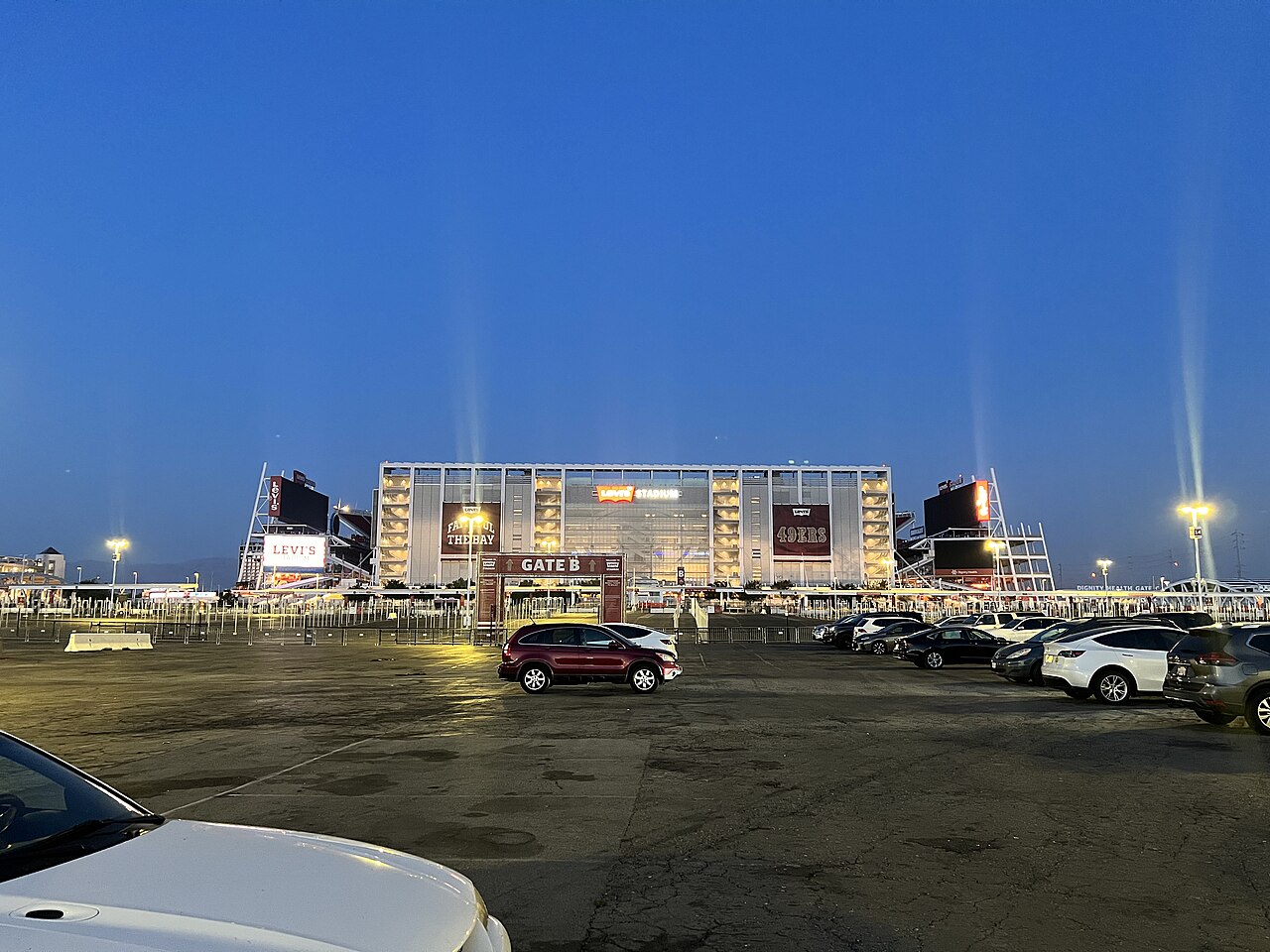 A brightly lit stadium entrance marked Gate B behind a parking lot at dusk