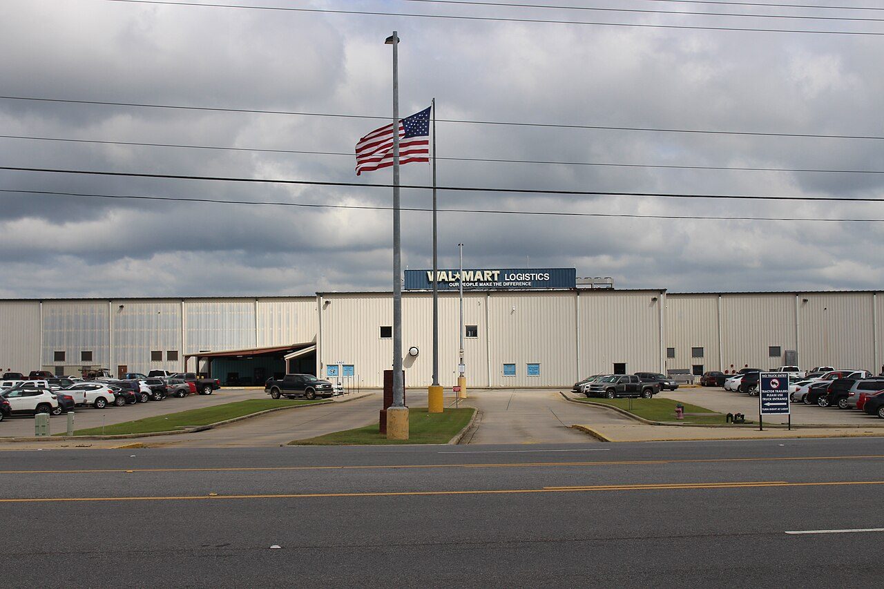 A large white warehouse with a Walmart sign, an American flag and a parking lot beside a road