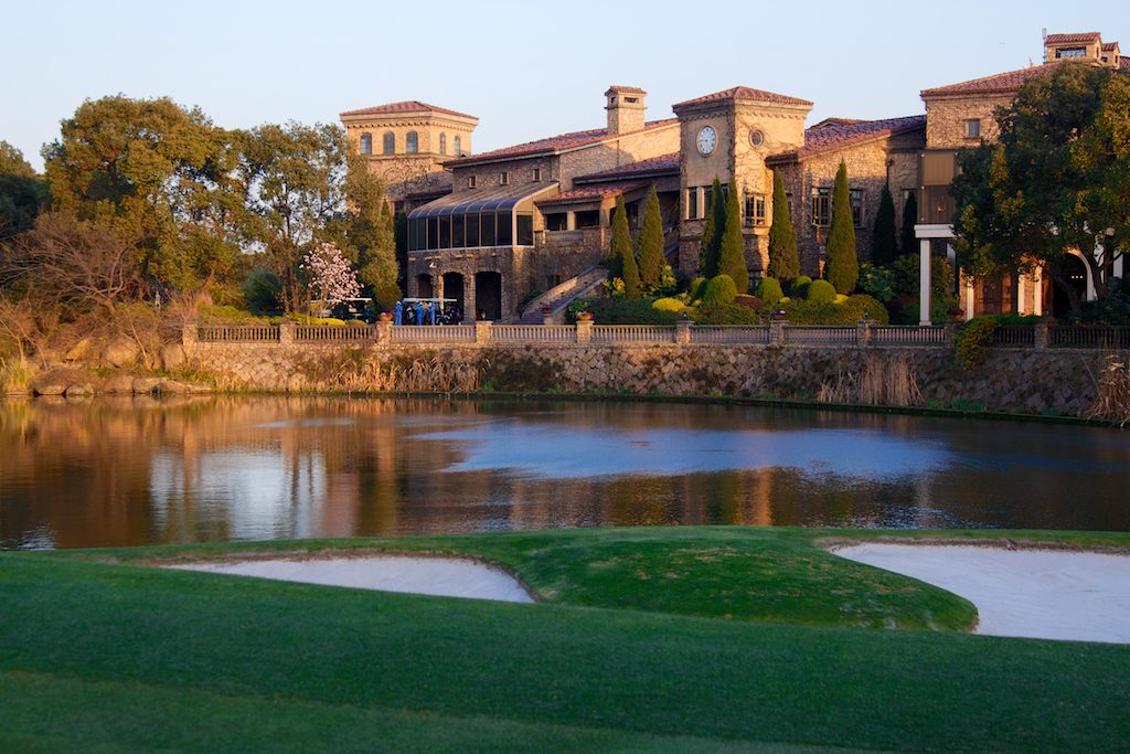 A golf green beside a pond, with a large Mediterranean-style clubhouse among trees in the background