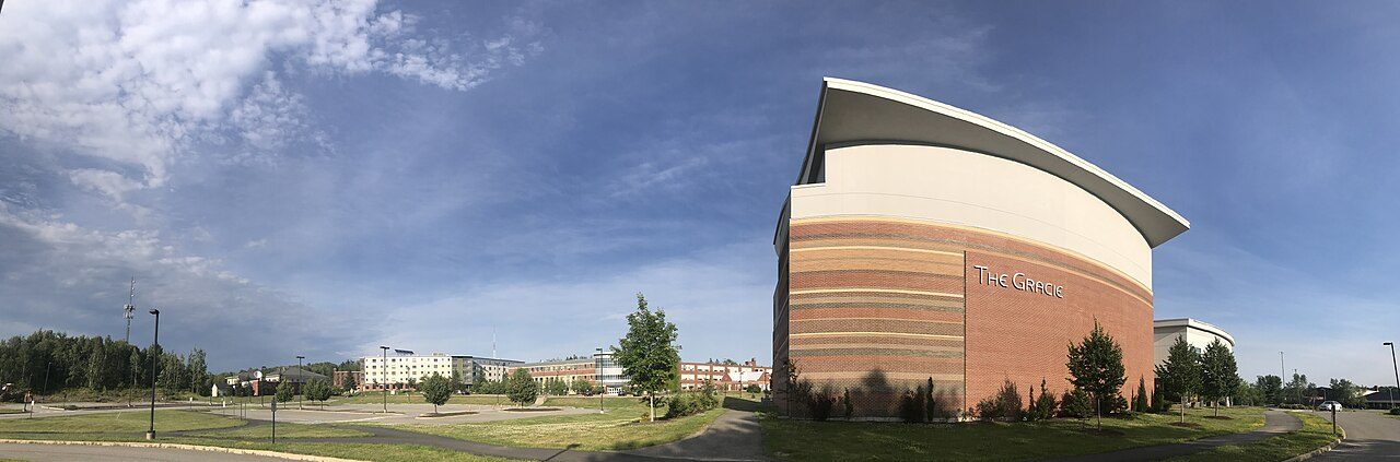 A modern brick and cream-colored theater building with a curved roof stands beside a wide lawn and parking area under a cloudy sky