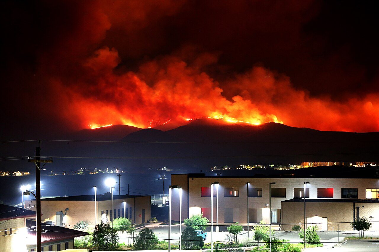A long wall of orange flames and red smoke glows along a dark ridge at night, above lit buildings and a parking area in the foreground