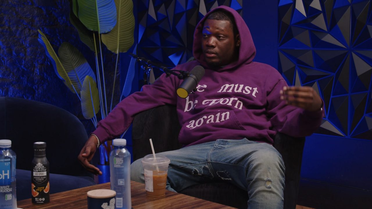 Michael Che, wearing a purple hooded sweatshirt and jeans, sits in a chair and talks into a microphone in a studio lit with blue light
