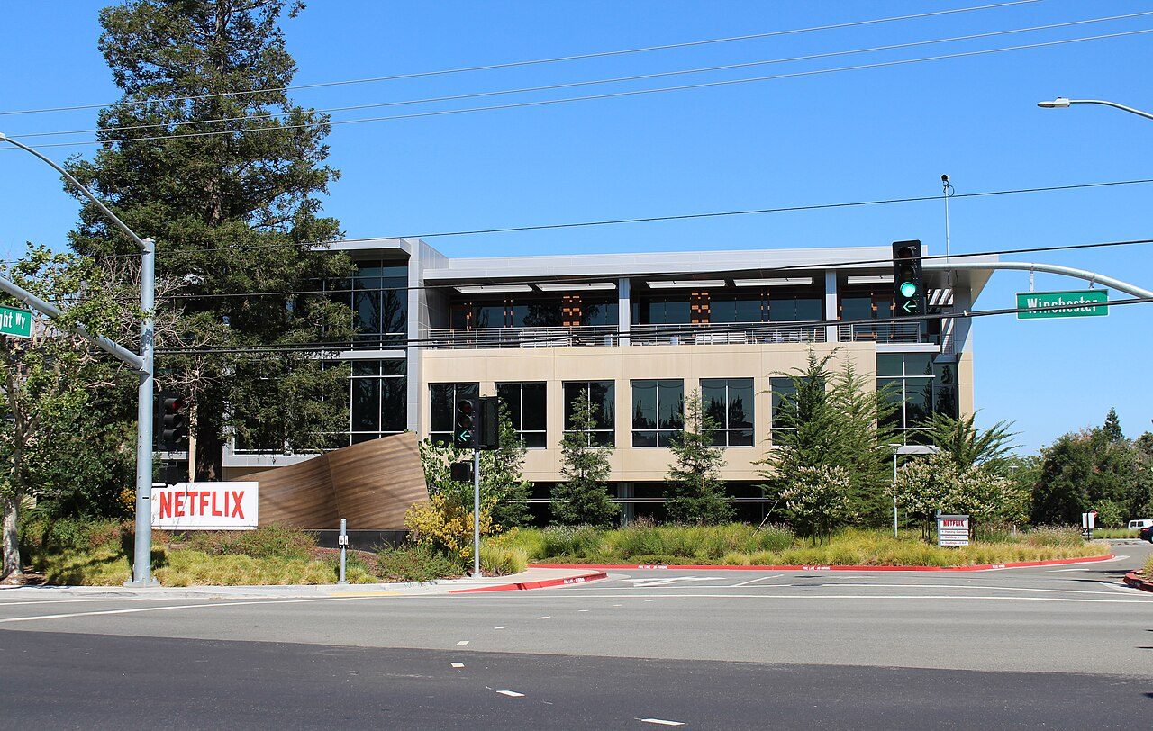 A low beige office building with a red Netflix sign out front, seen across a sunny intersection with trees and traffic lights