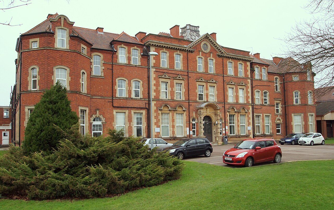 A red-brick Victorian hospital building with a lawn and parked cars in front