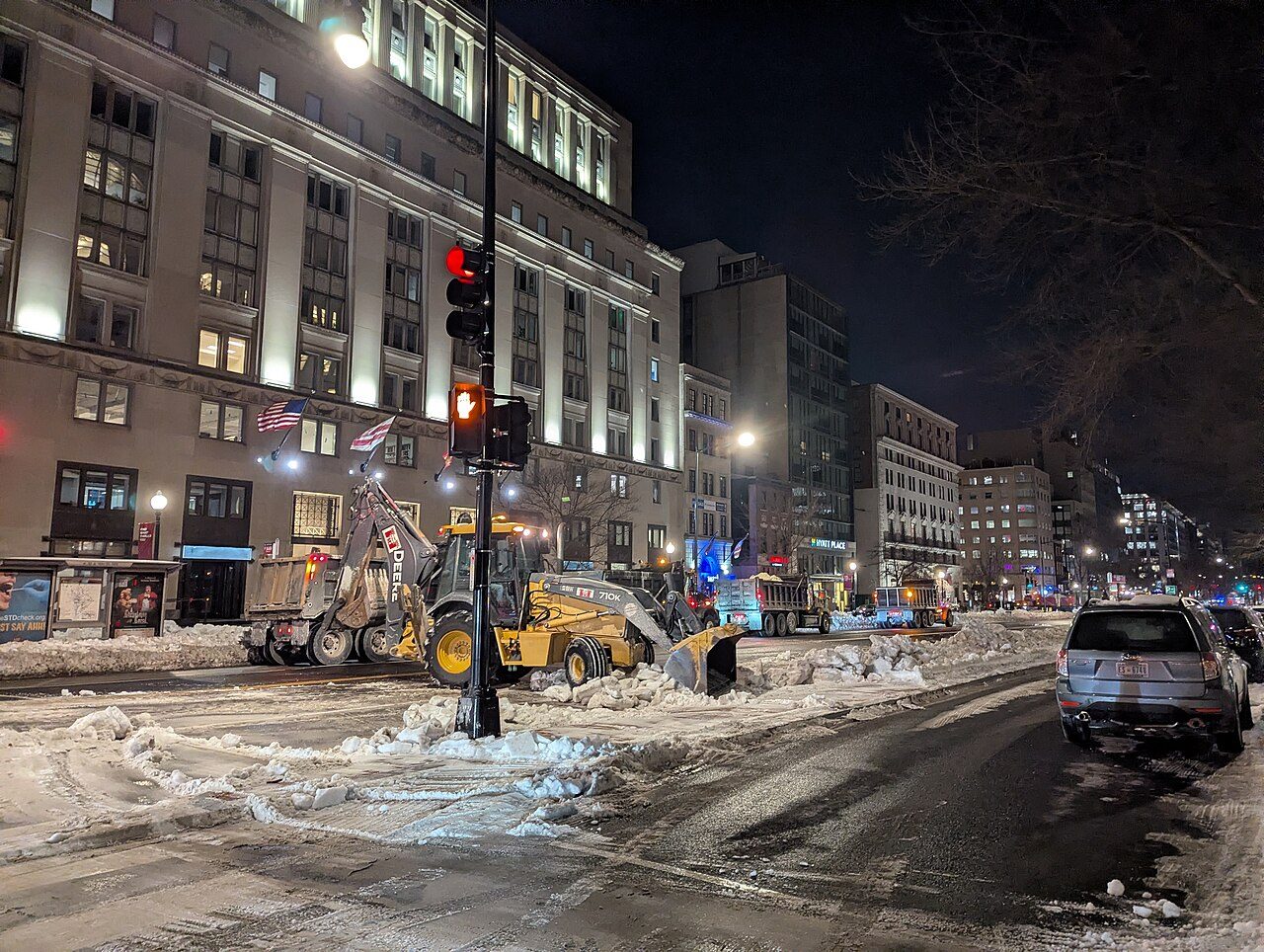 A backhoe pushes plowed snow at a city intersection at night, with a red traffic light, a lit office building and parked cars on a slushy street