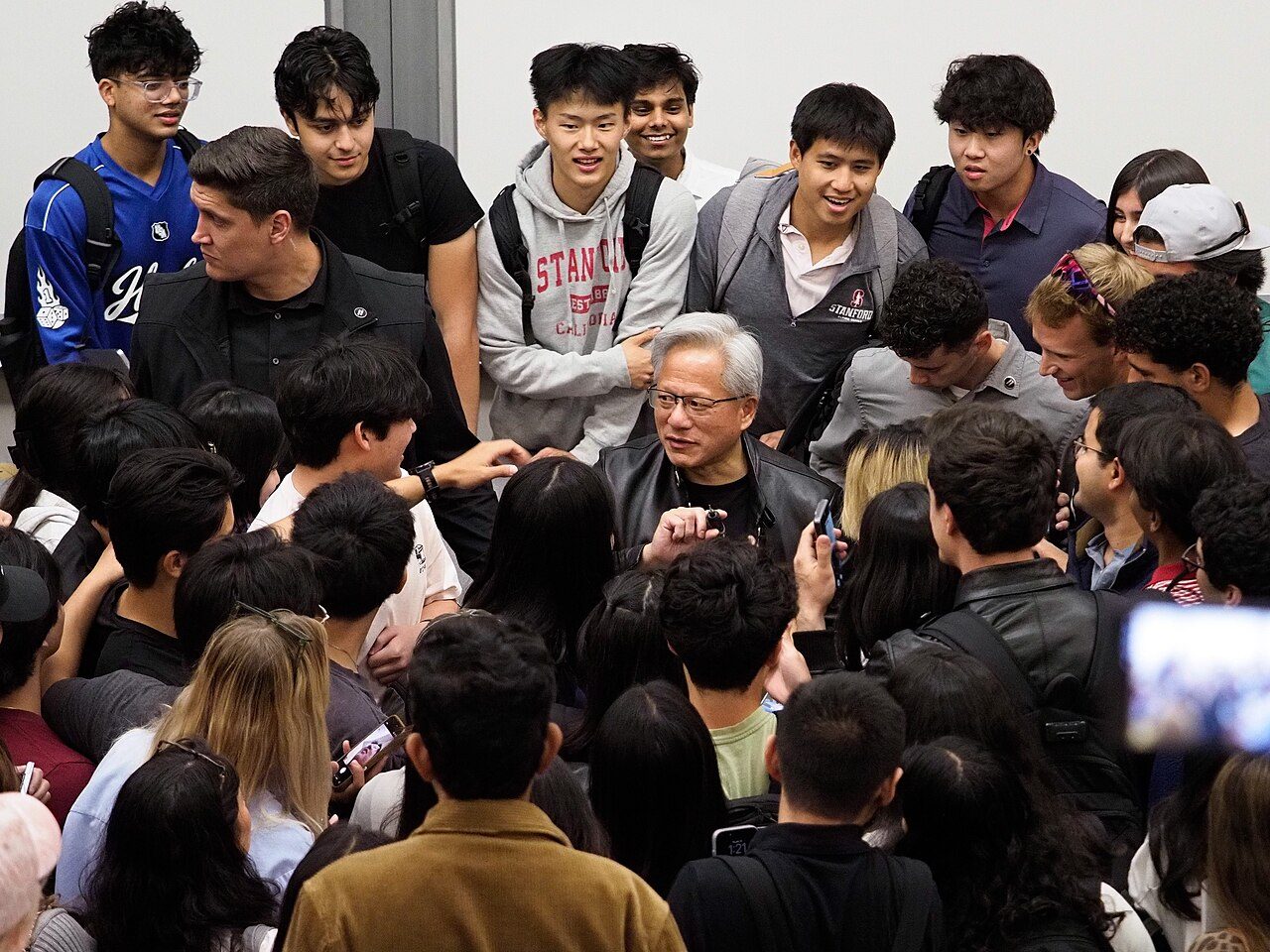 A gray-haired man in a dark jacket surrounded by a crowd of young people, some holding up phones
