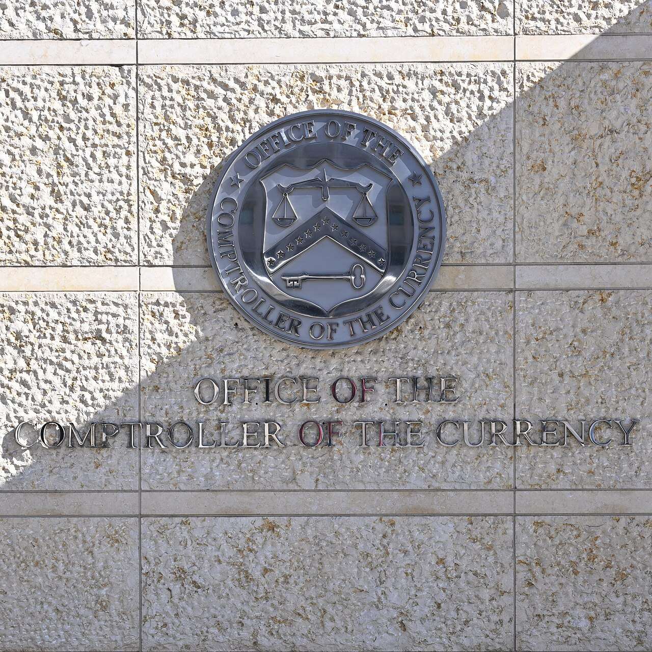 A round seal and the words Office of the Comptroller of the Currency on a light stone wall