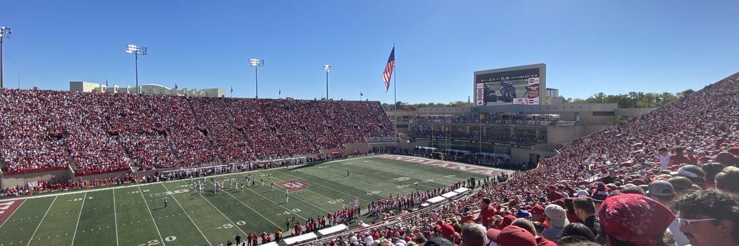 A packed college football stadium seen from the stands, with fans in red filling both sides and a green field below a blue sky