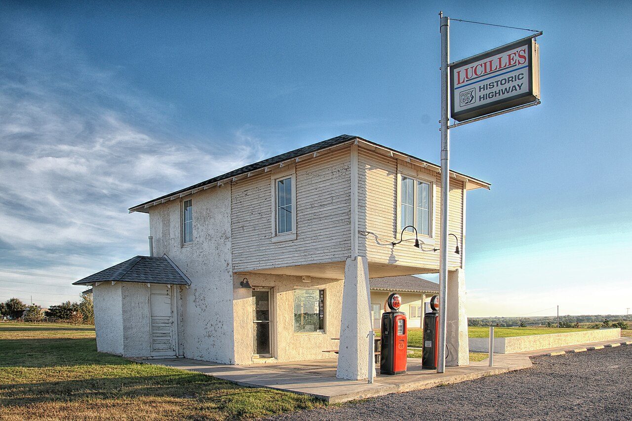 A small white two-story former gas station with a red pump out front and a tall sign, on a paved lot beside a grassy field under a blue sky
