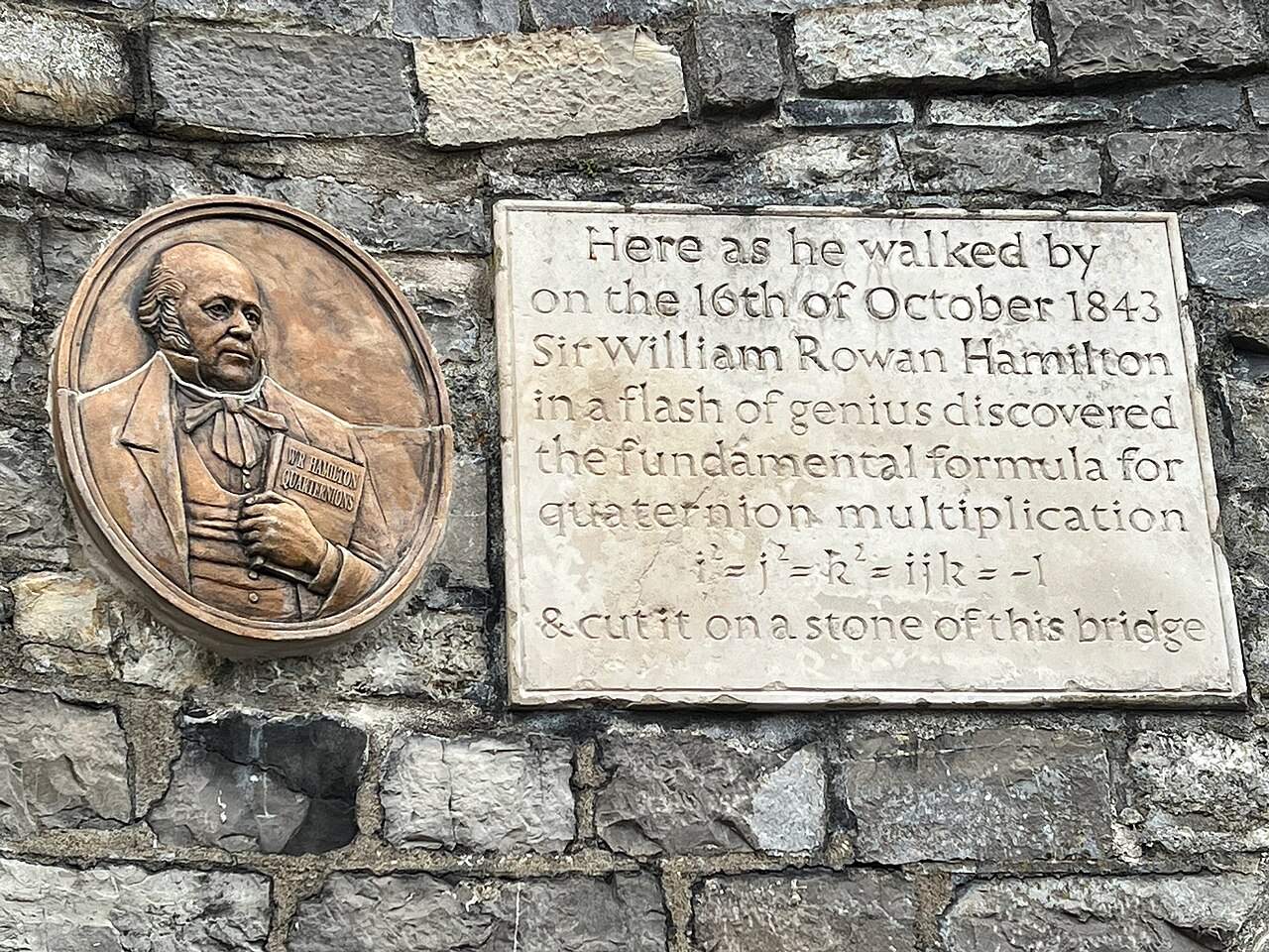A stone plaque on a rough stone wall reading that Sir William Rowan Hamilton discovered the formula for quaternion multiplication here on Oct. 16, 1843, beside a bronze medallion of Hamilton