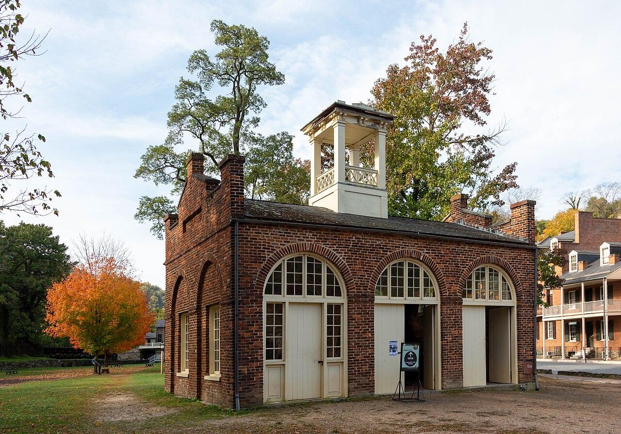 A small red-brick fire-engine house with arched doorways and a white cupola, with autumn trees behind it