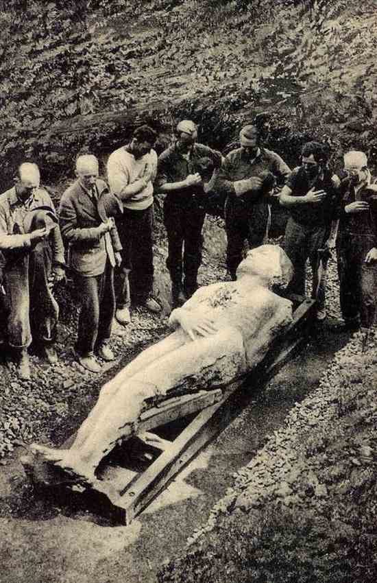 A black-and-white photo of a large pale stone figure of a man lying in a dug-out pit while a row of men stand around it with their hats off