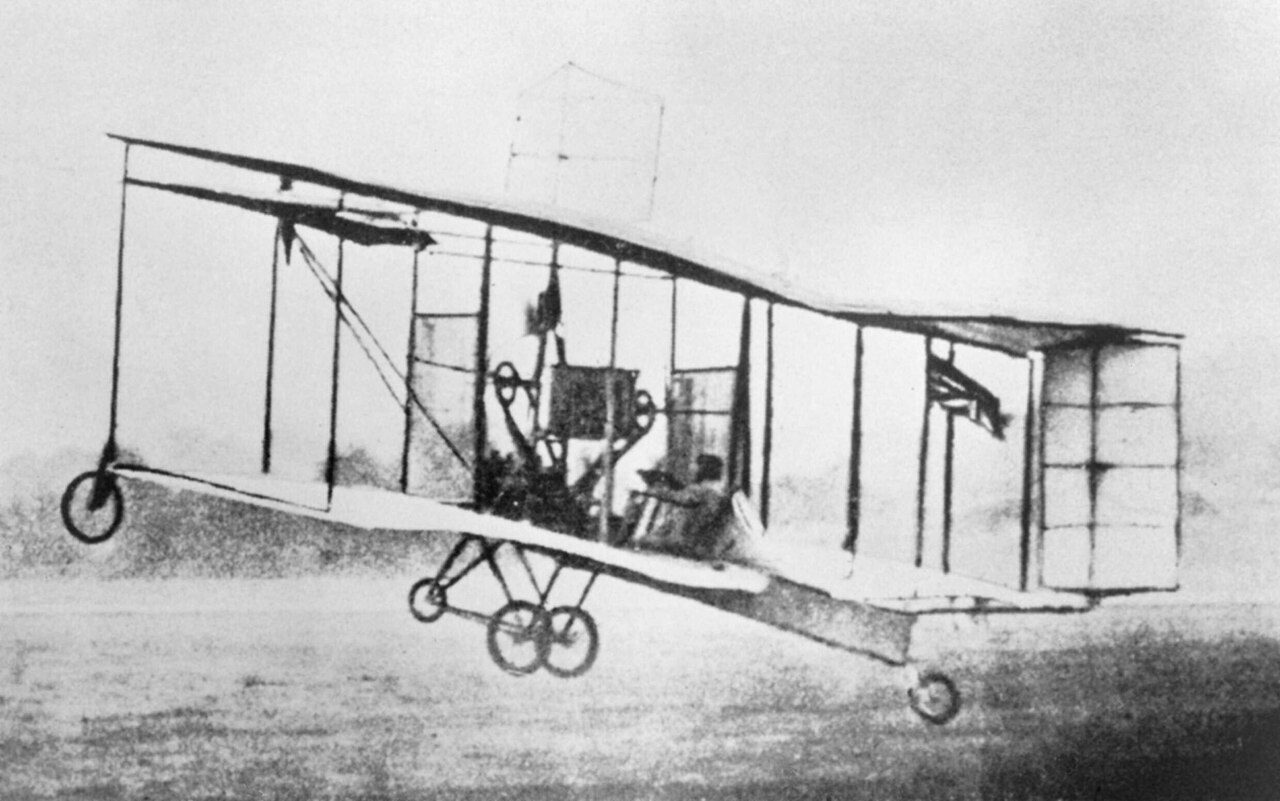 A black-and-white photo of an early biplane with large box-shaped wings and a front elevator flying low over a field