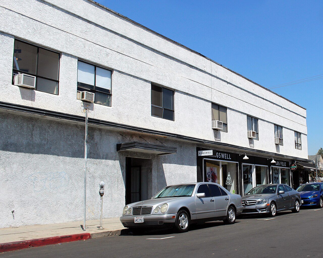 A plain two-story white commercial building on a sunny street corner, with cars parked along the curb