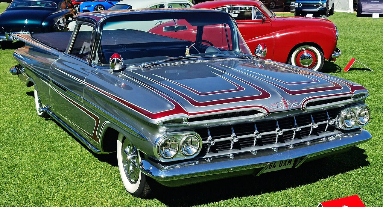 A silver 1959 Chevrolet El Camino with red pinstriping, a wide chrome grille and twin headlights, parked on grass at a car show