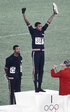 An athlete wearing number 307 stands on the top step of an Olympic podium with both arms raised, while another athlete stands on a lower step