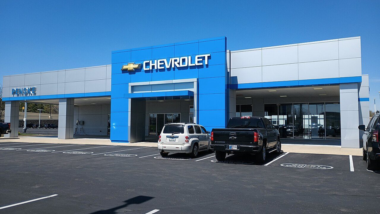 A car dealership with a blue and white glass front and a large Chevrolet sign, with pickup trucks and cars parked out front under a clear sky