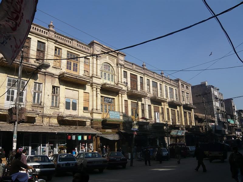A row of old multistory buildings with balconies and shop signs lines a busy street in Peshawar's Old City, with parked cars and overhead power lines