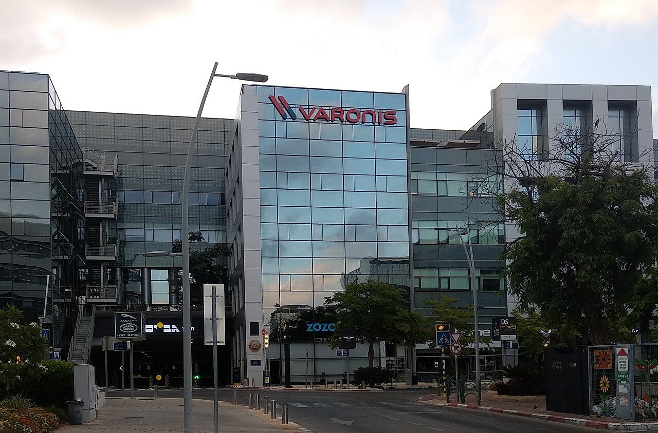 A modern glass office building with a red Varonis sign on its upper floor, next to a parking lot and trees