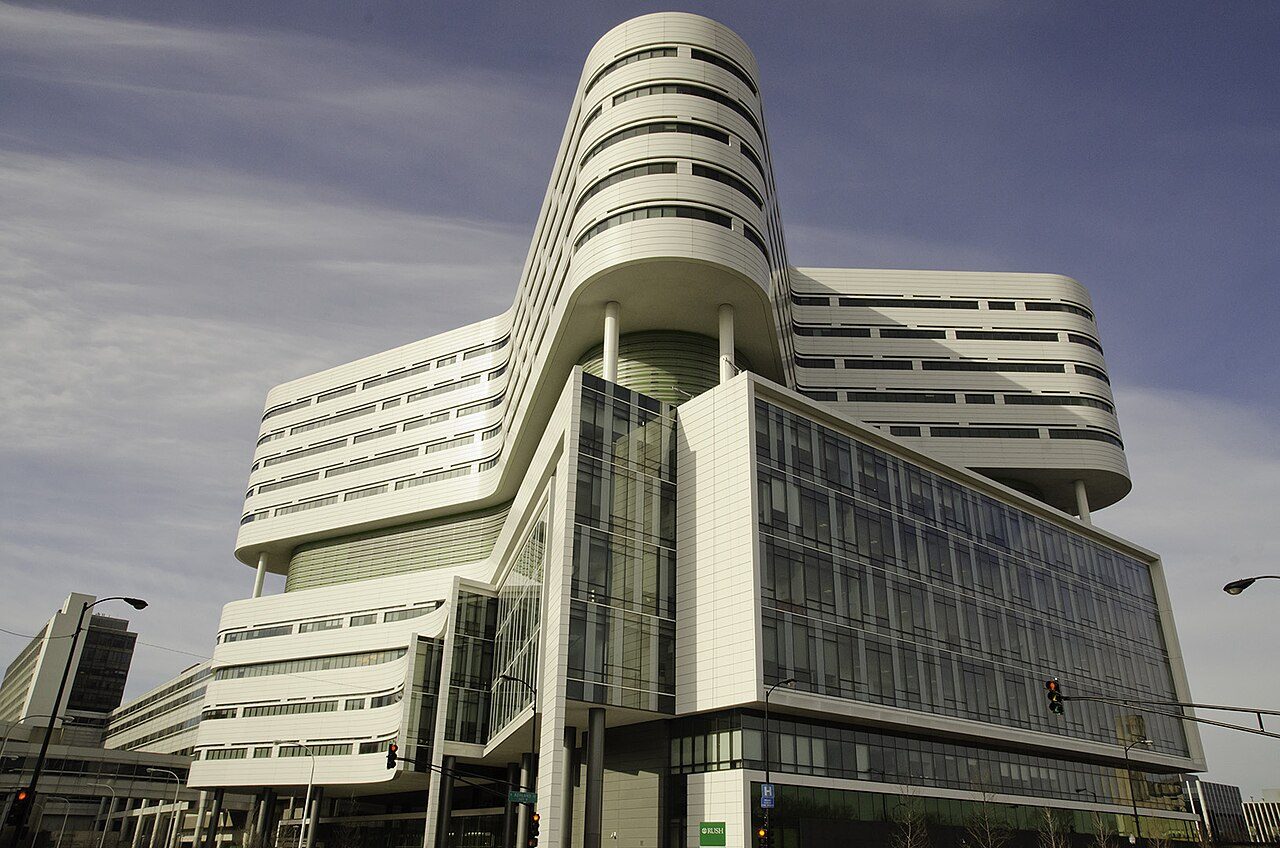 A tall white hospital tower with curving, stacked floors and glass walls rises above a street corner under a cloudy blue sky
