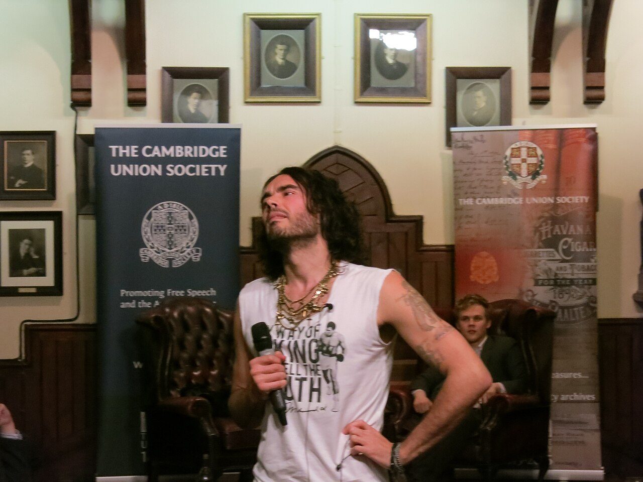 Russell Brand holding a microphone in a wood-paneled room with Cambridge Union Society banners
