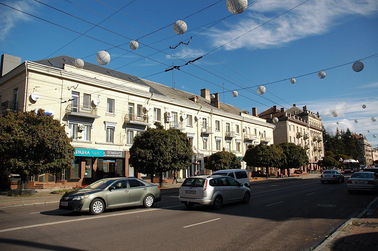 A sunny street in Lutsk lined with cream-colored historic buildings and trees, with cars on the road and round lamps strung on wires overhead