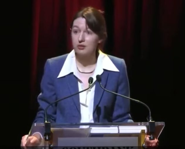 Sally Rooney in a blue jacket speaking at a lectern with microphones