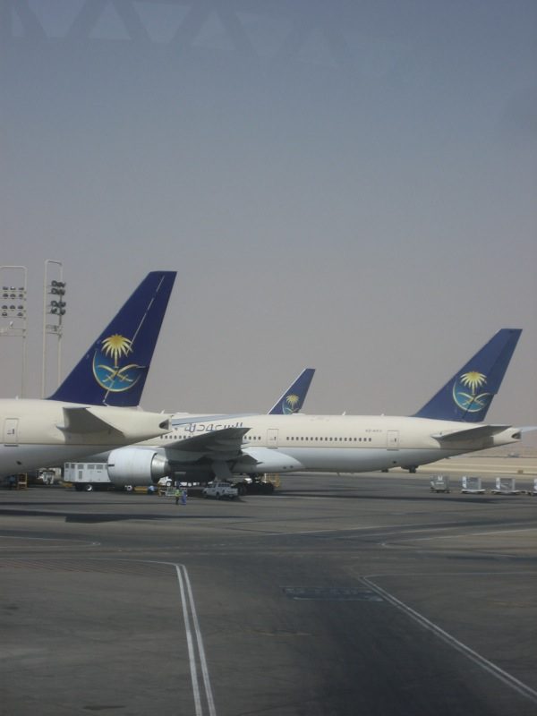 Several Saudi Arabian Airlines jets with blue tails and palm-and-swords logos parked at an airport terminal apron under a hazy sky