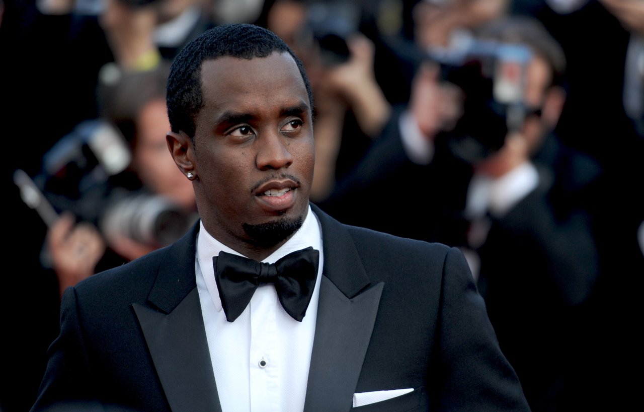 Sean Combs, in a black tuxedo and bow tie, stands in front of a crowd of photographers on a red carpet