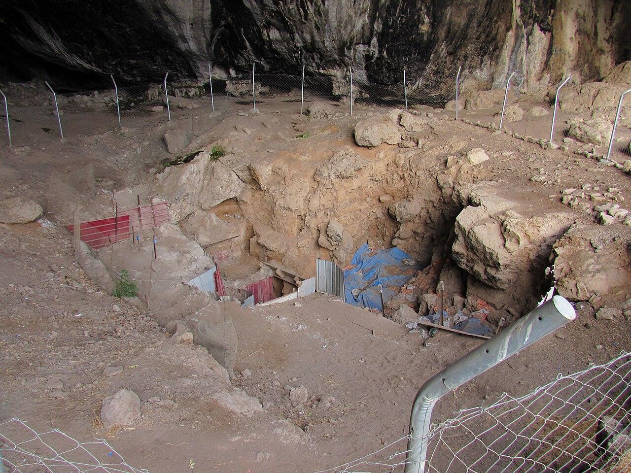 Inside a rocky cave, a deep excavation pit in pale dirt and boulders, with a blue tarp at the bottom and a metal fence along one side