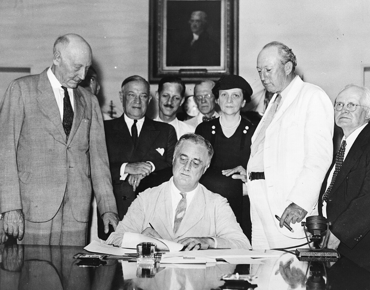 Black-and-white photo of a seated man signing a document while a group of men and a woman stand behind him