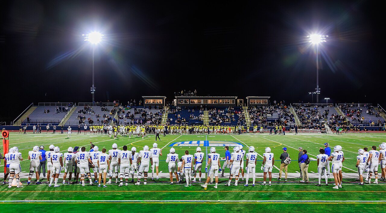 Football players in white and blue uniforms stand along the sideline of a lit stadium at night, with fans in the stands behind them