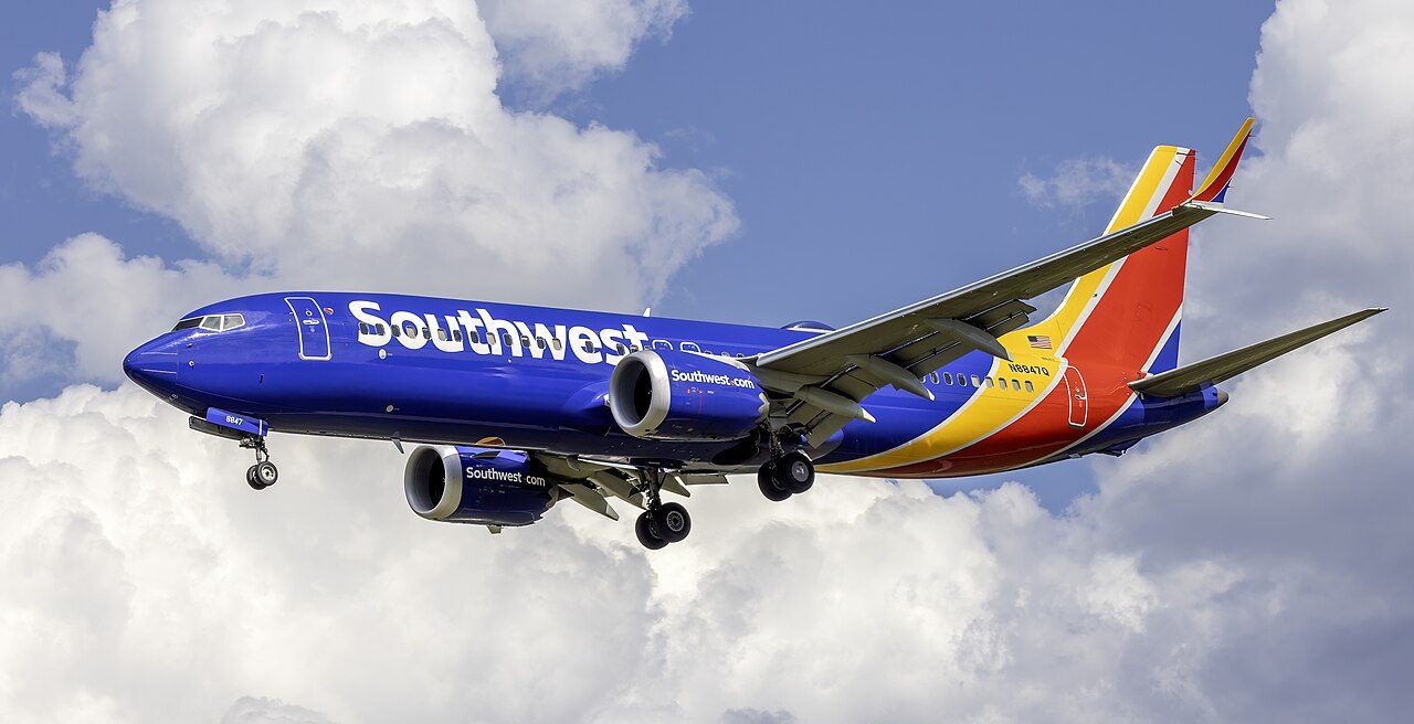 A blue Southwest Airlines Boeing 737 with red and yellow tail stripes flies with its landing gear down against white clouds and blue sky