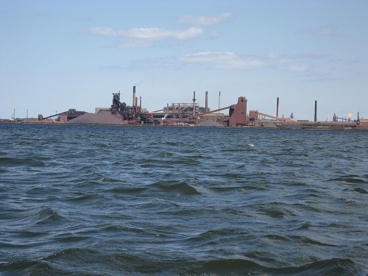 A steel plant with smokestacks on the far shore of choppy blue water
