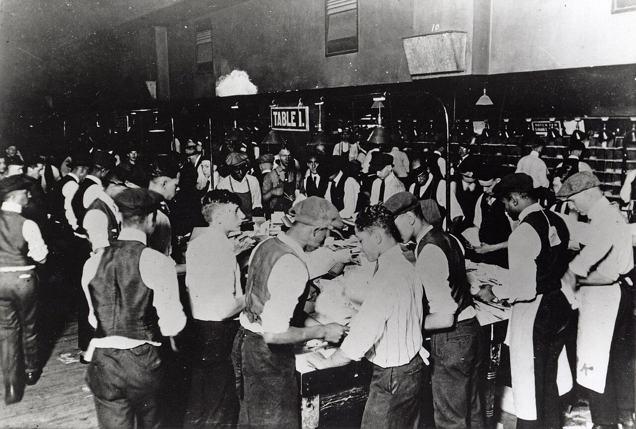 Black-and-white archival photo of a crowded post office floor where dozens of clerks in shirtsleeves and aprons sort letters by hand at long tables