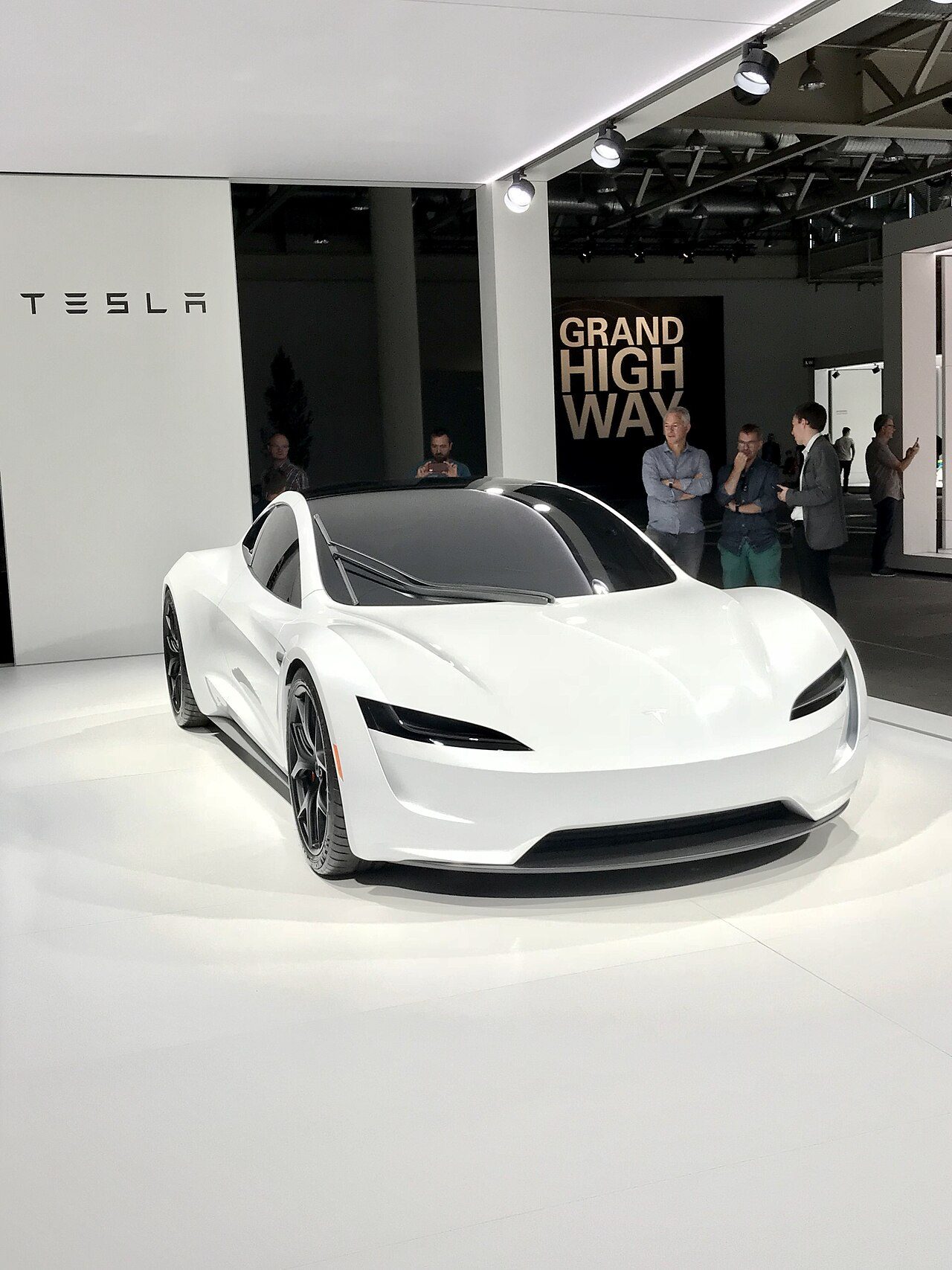 A white two-door Tesla Roadster sports car on a white stage under bright lights, with a Tesla sign on the wall behind it