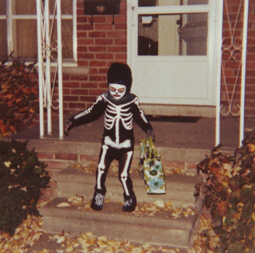 A child in a black skeleton costume and mask walks down the brick front steps of a house holding a treat bag, with fallen leaves on the ground