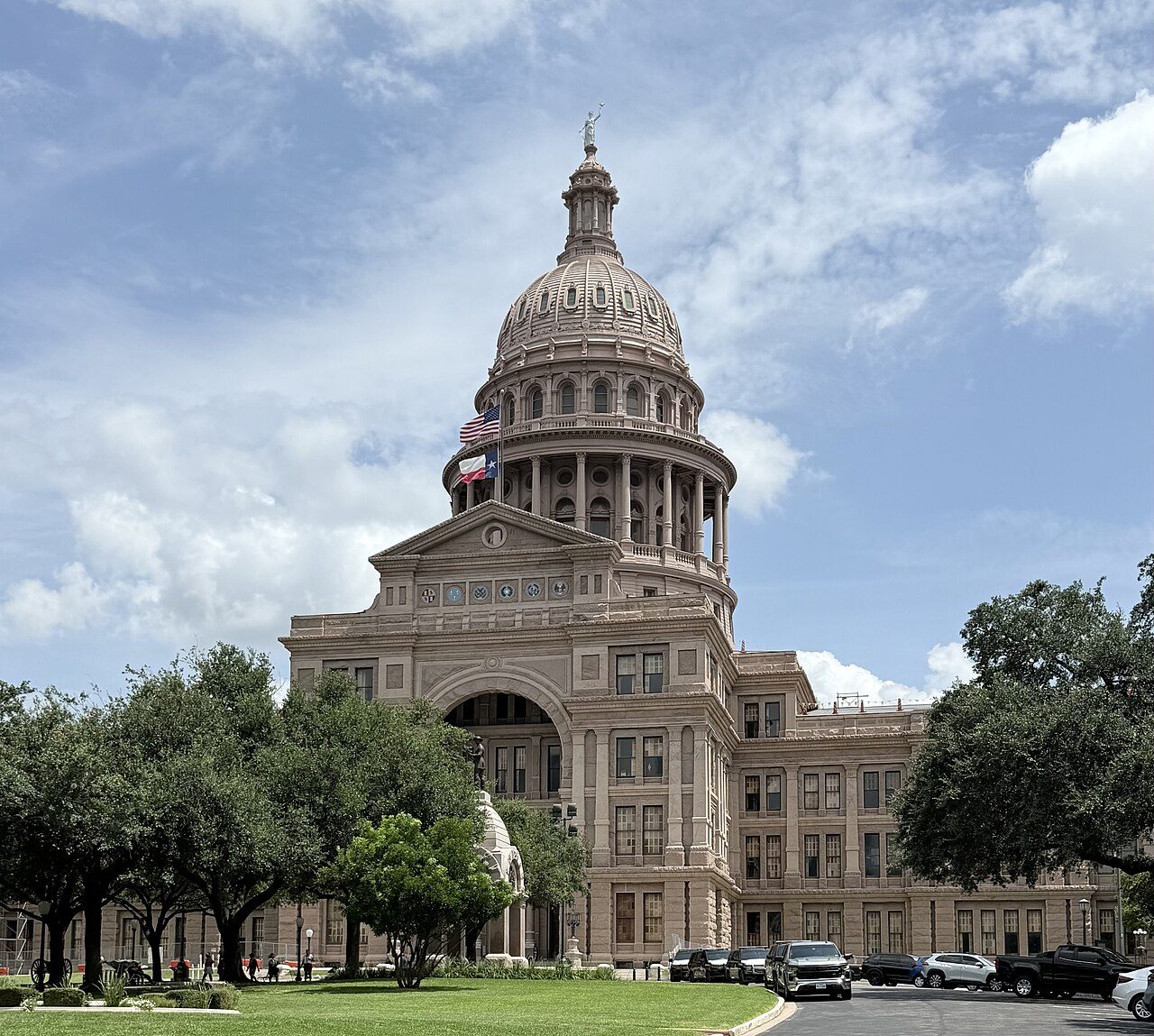 The Texas State Capitol's pink granite dome and front facade under a partly cloudy sky, framed by green trees and a lawn