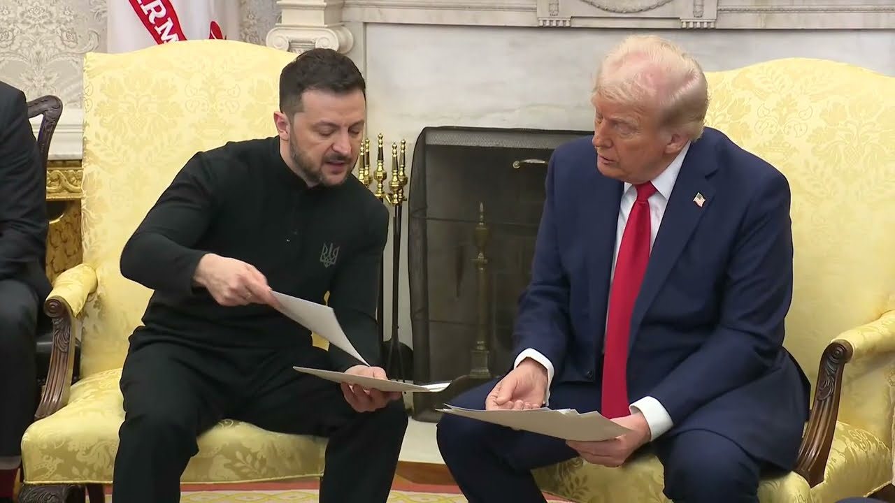 Ukrainian President Volodymyr Zelenskyy, dressed in black, hands papers to President Donald Trump, in a blue suit and red tie, as they sit in yellow armchairs by a fireplace