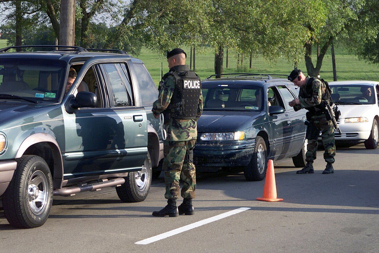 Soldiers in camouflage and helmets check cars lined up at a base entrance, with an orange traffic cone on the road and trees behind
