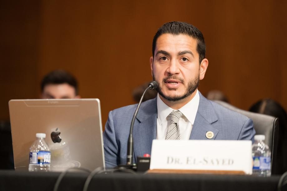 A man in a suit speaks into a microphone at a hearing table with a name placard in front of him