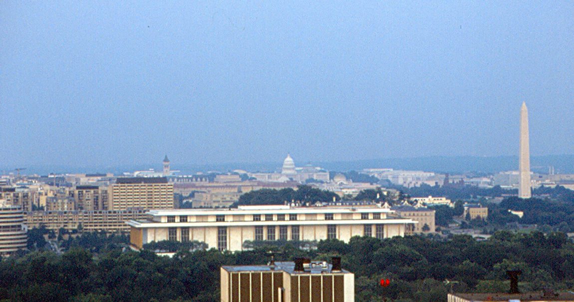 A long white building with a flat roof sits among trees in the foreground, with the Washington Monument, the U.S. Capitol dome and city buildings behind it under a hazy sky