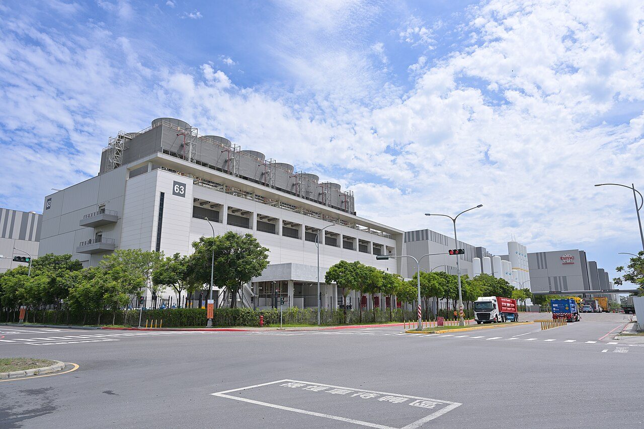 A long, low white TSMC factory building with rows of windows behind a wide, empty road and trees under a blue sky with scattered clouds
