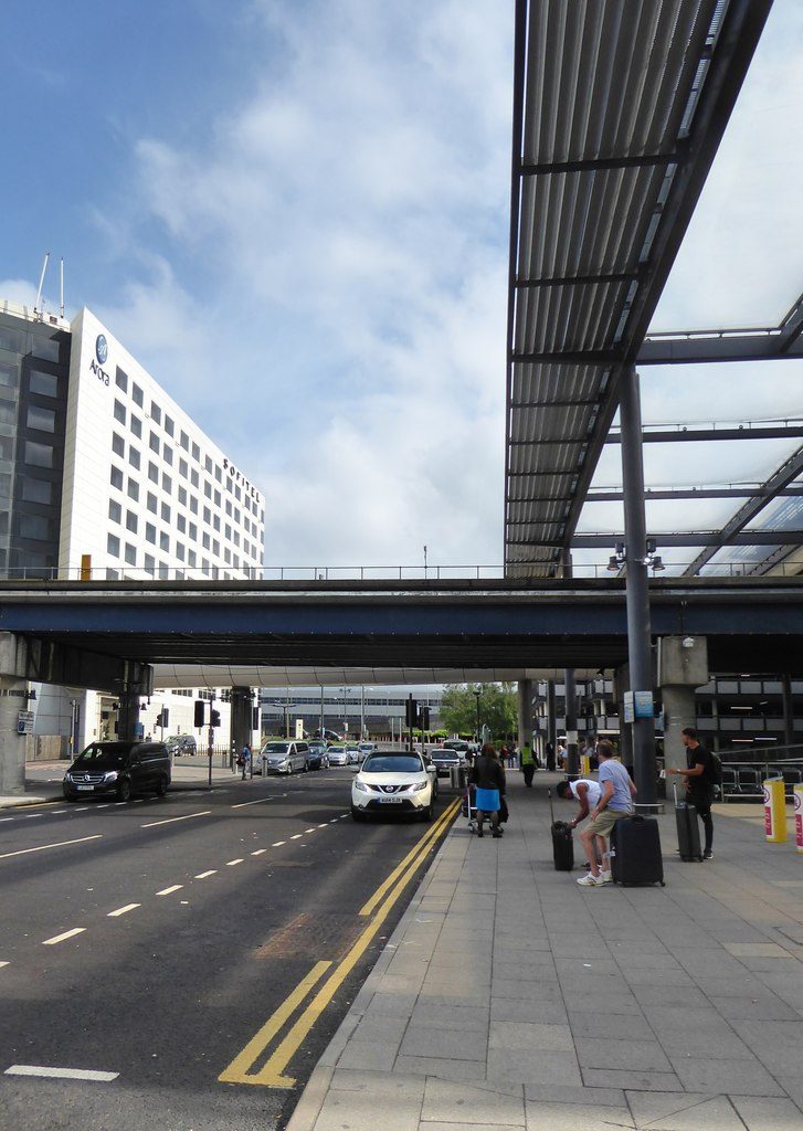 A covered drop-off lane outside an airport terminal, with a few travelers on the sidewalk and cars in the road